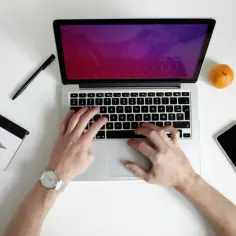 Ręce piszą na laptopie, obok notatnik, długopis, mandarynka i telefon. Idealne do pracy zdalnej, gdy liczy się czas pracy baterii.