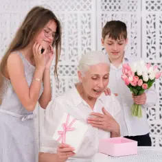 Wzruszona babcia otwiera prezent na Dzień Babci i Dziadka. Dzieci wręczają jej bukiet tulipanów i pudełko z upominkiem.