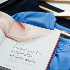 Książka "Encyklopedia materiałów odzieżowych" obok błękitnej i granatowej bluzki na wieszakach. Dowiedz się, jak czytać etykiety ubrań, żeby rozpoznać poliester od wiskozy.