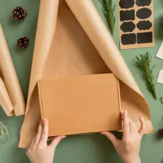 Ręce owijają kartonowe pudełko brązowym papierem. Wokół leżą szyszki, gałązki sosny, sznurek i nożyczki – idealne do pakowania prezentów.
