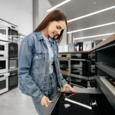 Kobieta w dżinsowej kurtce ogląda płytę indukcyjną, zastanawiając się nad różnicą między produktem outletowym a produktem powystawowym.