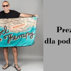 Uśmiechnięty mężczyzna w okularach prezentuje ręcznik z mapą świata i napisem "Travel is my therapy". Idealny prezent dla podróżnika.