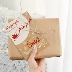 Ręka trzyma paczkę z papieru pakowego, ozdobioną kartką z Mikołajem i reniferami. To z pewnością fajne prezenty na święta!