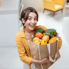 Uśmiechnięta kobieta z torbą pełną świeżych owoców i warzyw. Dobry wybór produktów to klucz do zdrowia i satysfakcji, unikaj skutków kupowania produktów z dołu rankingu opłacalność vs jakość.