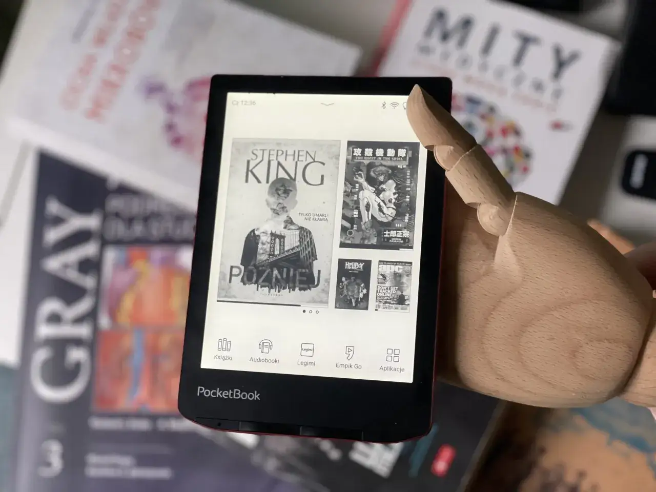 Drewniana dłoń wskazuje na ekran czytnika PocketBook z okładkami książek. To idealny gadżet dla miłośników czytania, oferujący najlepsze czytniki e-booków ranking modeli z podświetleniem ekranu.