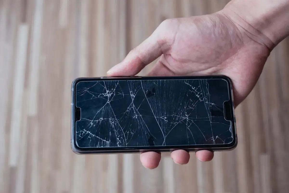 Dłoń trzyma smartfon z pękniętym ekranem. To wyraźne objawy uszkodzonego wyświetlacza w używanym smartfonie przed zakupem.