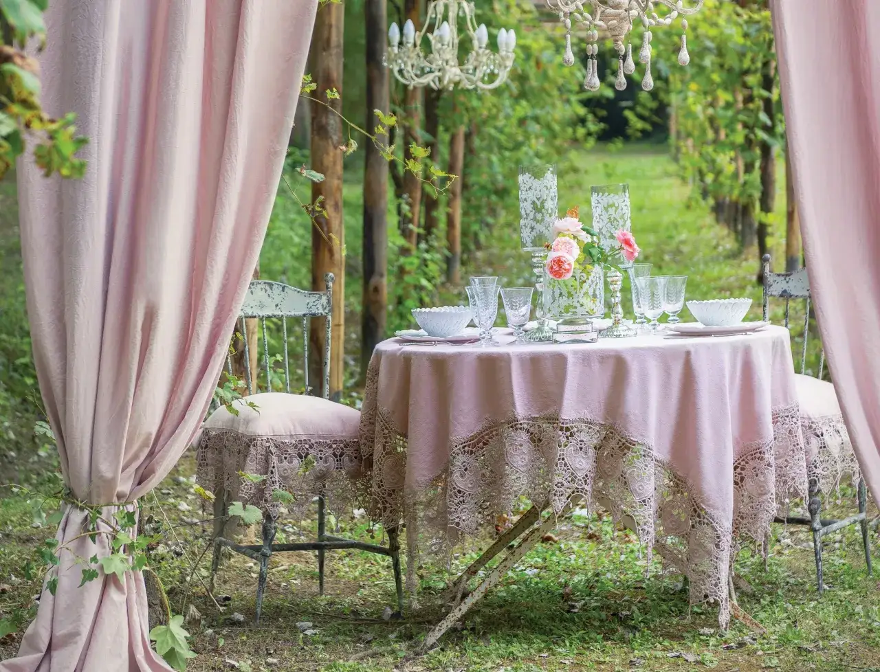 Stół na przyjęcie w ogrodzie w stylu boho: różowy obrus z koronką, kryształowy żyrandol, kwiaty i pastelowe zasłony tworzą magiczną atmosferę.
