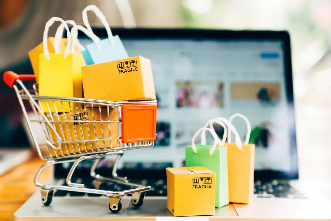 Miniaturowy wózek z zakupami i paczkami na laptopie ilustruje różnicę między handlem tradycyjnym a e-commerce, koszty i marże.